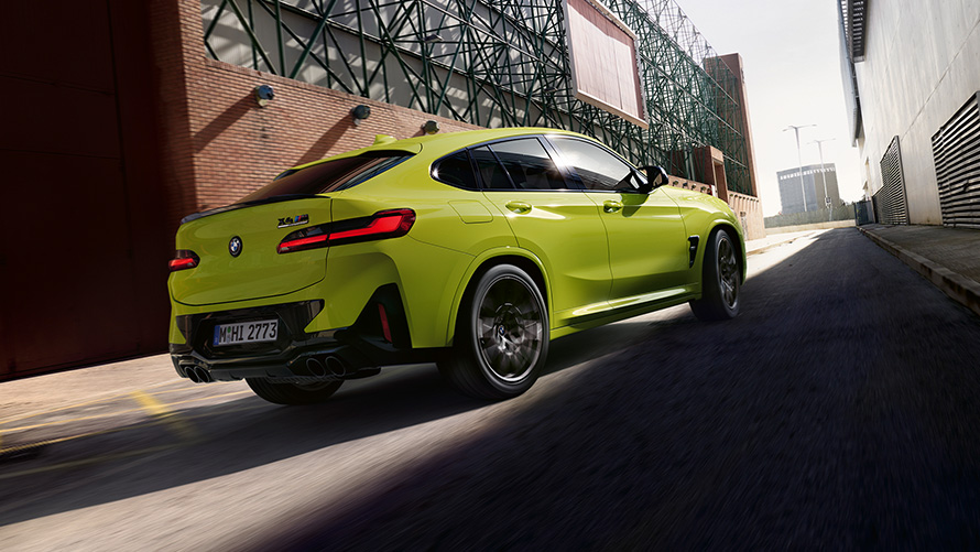 BMW X4 M Competition F98 LCI Facelift 2021 v barvě Sao Paulo Yellow (žlutá) z tříčtvrtečního pohledu zepředu při jízdě mezi továrními budovami